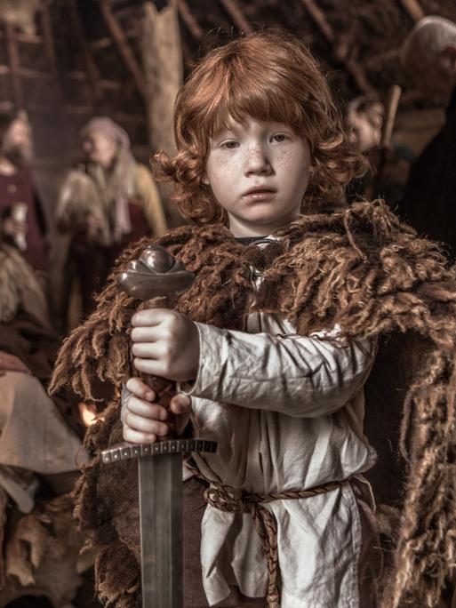 Eine Wikingerfamilie, im Vordergrund ein rothaariger Junge im Fellumhang mit Waffe