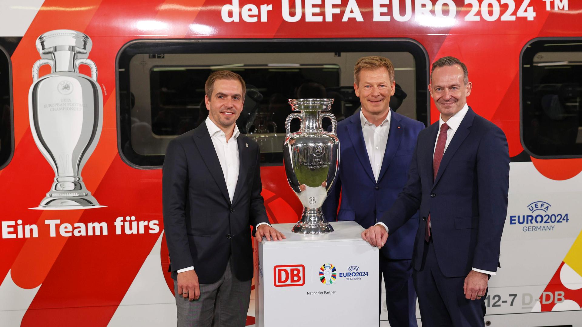 EM-Botschafter Philipp Lahm, DB-Chef Richard Lutz und Volker Wissing, Bundesverkehrsminister, stehen bei der Bekanntgabe der Partnerschaft zwischen Deutscher Bahn und der UEFA zur Europameisterschaft 2024 vor dem EM-Pokal und einem ICE.