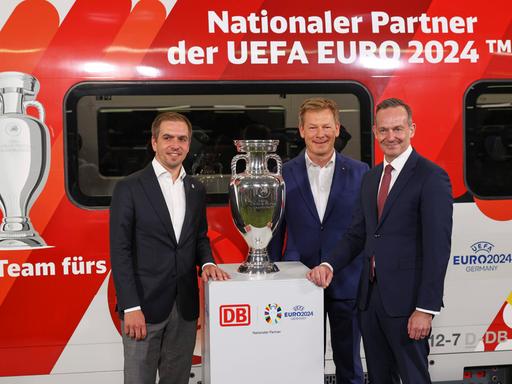 EM-Botschafter Philipp Lahm, DB-Chef Richard Lutz und Volker Wissing, Bundesverkehrsminister, stehen bei der Bekanntgabe der Partnerschaft zwischen Deutscher Bahn und der UEFA zur Europameisterschaft 2024 vor dem EM-Pokal und einem ICE.