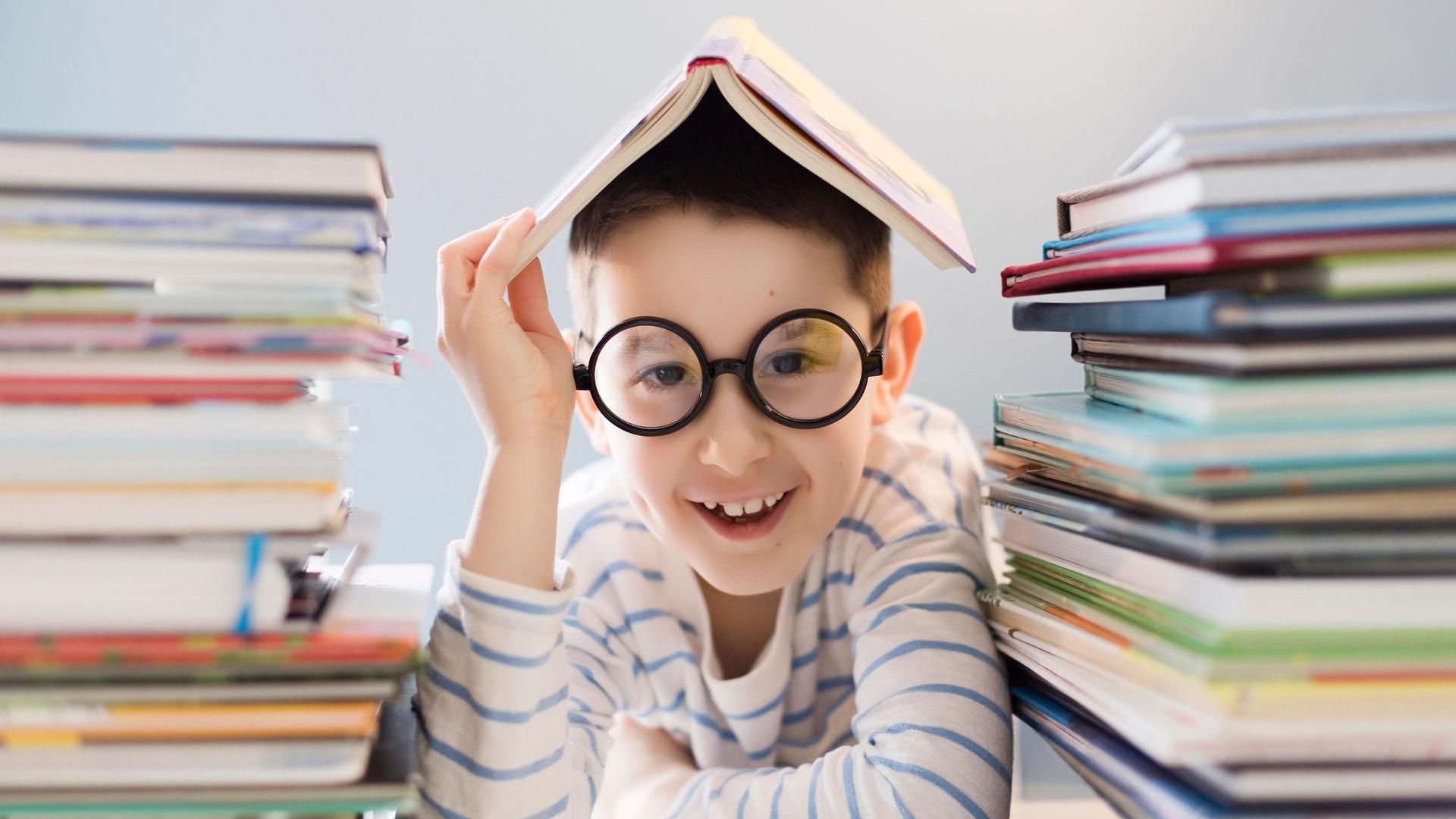 Ein Junge sitzt zwischen 2 Bücherstapekn mit einem Buch auf dem Kopf