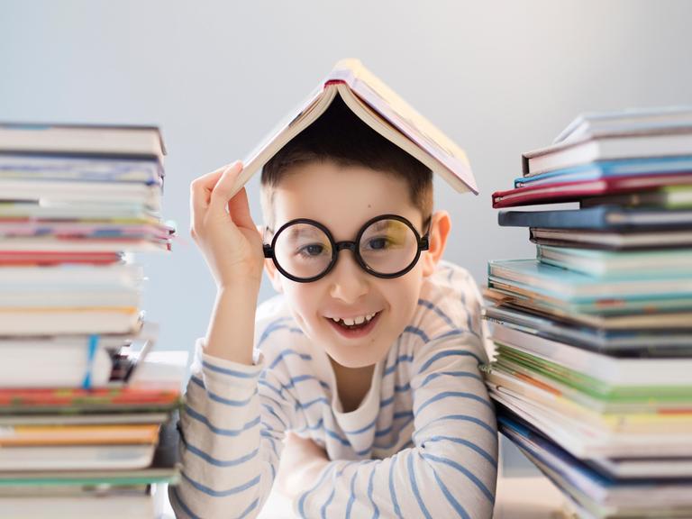 Ein Junge sitzt zwischen 2 Bücherstapekn mit einem Buch auf dem Kopf