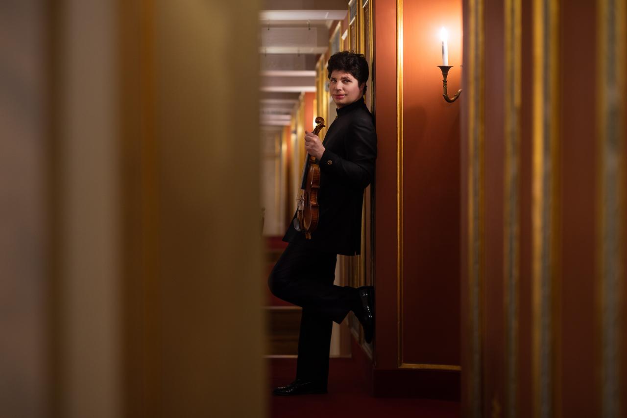 Ein junger Mann mit Geige lehnt im Gang eines Ranges des Konzerthauses Berlin an der Wand mit Wandleuchter und blickt seitlich in die Kamera.
