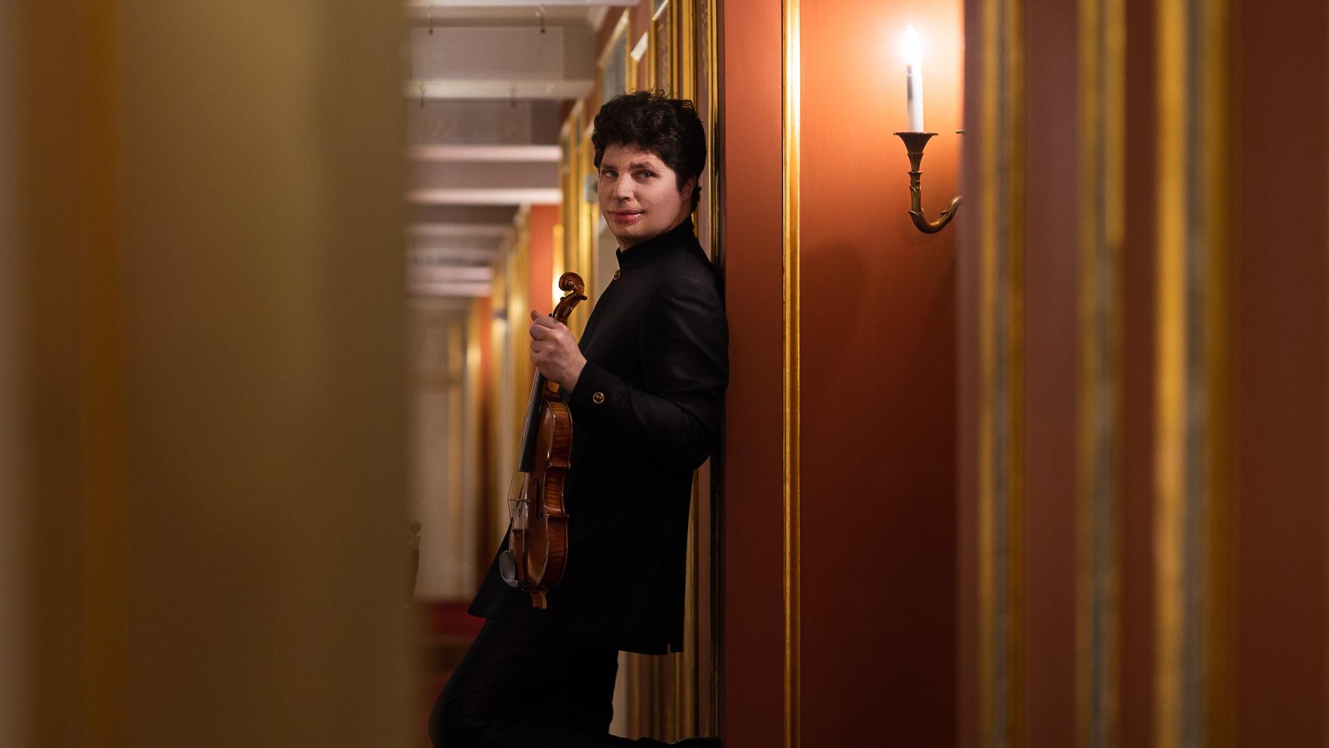 Ein junger Mann mit Geige lehnt im Gang eines Ranges des Konzerthauses Berlin an der Wand mit Wandleuchter und blickt seitlich in die Kamera.