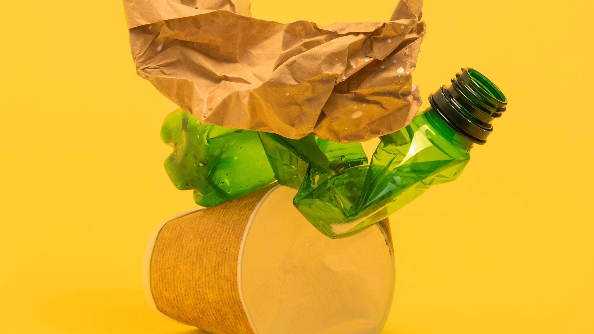Studioaufnahme von schwer zu recycelnden Kunststoffverpackungen wie Kaffebecher und folierte Verpackungen vor gelbem Hintergrund.