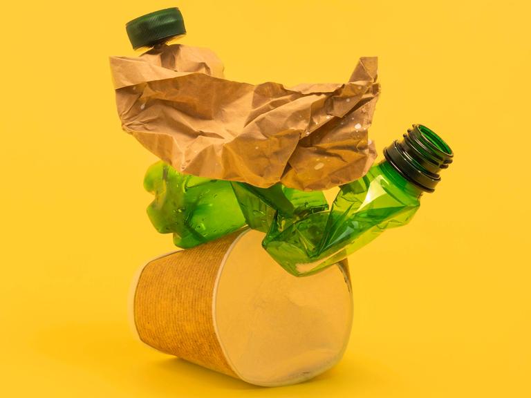 Studioaufnahme von schwer zu recycelnden Kunststoffverpackungen wie Kaffebecher und folierte Verpackungen vor gelbem Hintergrund.