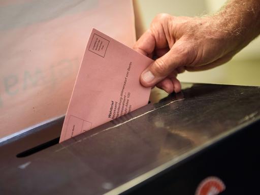 Eine männliche Hand steckt einen rosafarbenen Umschlag in den Schlitz einer Wahlurne.