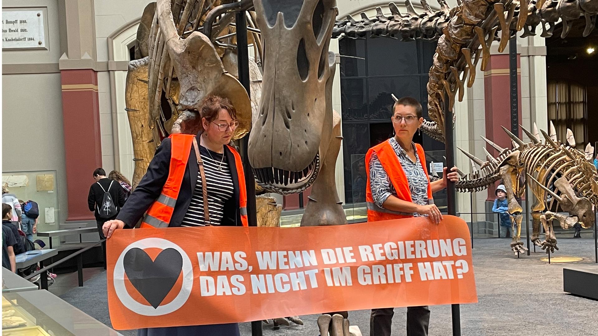 Zwei Klimaaktivistinnen mit einem Banner "Was, wenn die Regierung das nicht im Griff hat?" an den Haltestangen eines Dinosauriers festgeklebt. 