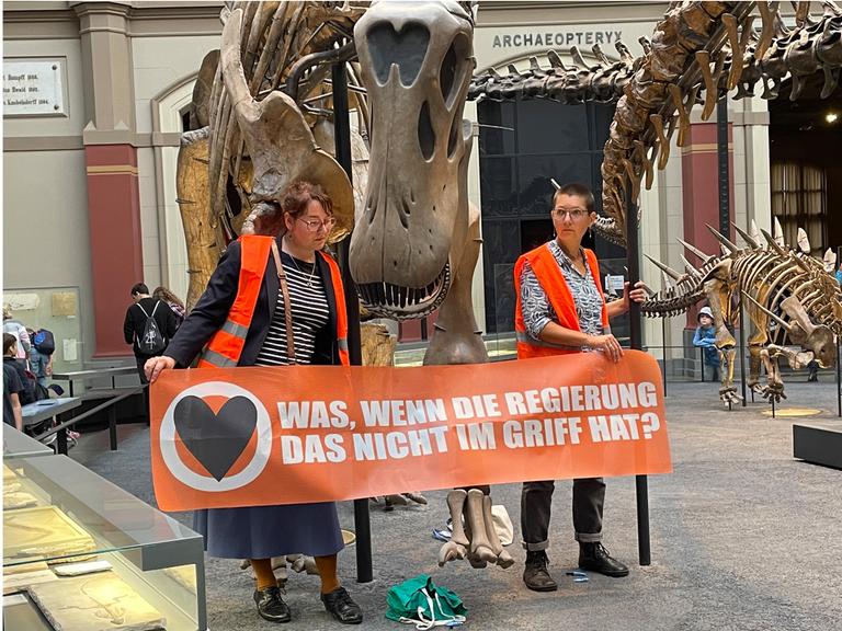 Zwei Klimaaktivistinnen mit einem Banner "Was, wenn die Regierung das nicht im Griff hat?" an den Haltestangen eines Dinosauriers festgeklebt. 