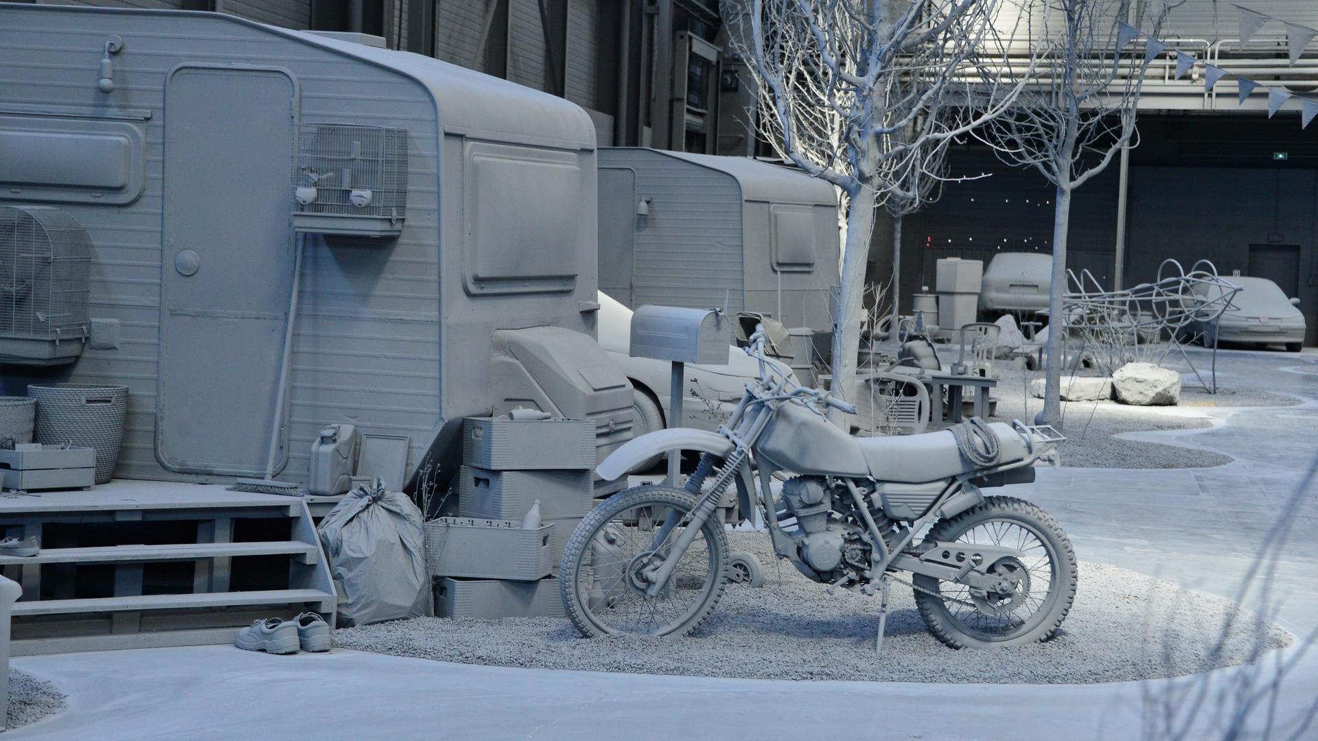 Eine Kunstinstallation zeigt ein Motorrad, einen Wohnwagen und andere Gegenstände in einem Hangar, in dem alles grau ist.