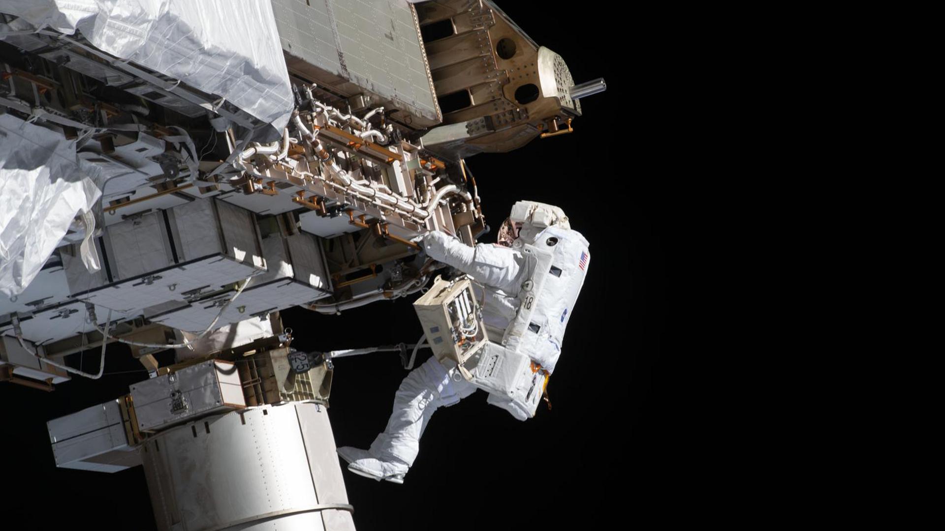 Ein Astronaut in einem weißen Raumanzug schwebt außerhalb der Internationalen Raumstation. Er hält sich mit beiden Händen an einer Metallstruktur fest und arbeitet an technischen Bauteilen.