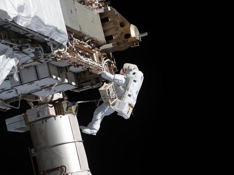 Ein Astronaut in einem weißen Raumanzug schwebt außerhalb der Internationalen Raumstation. Er hält sich mit beiden Händen an einer Metallstruktur fest und arbeitet an technischen Bauteilen.