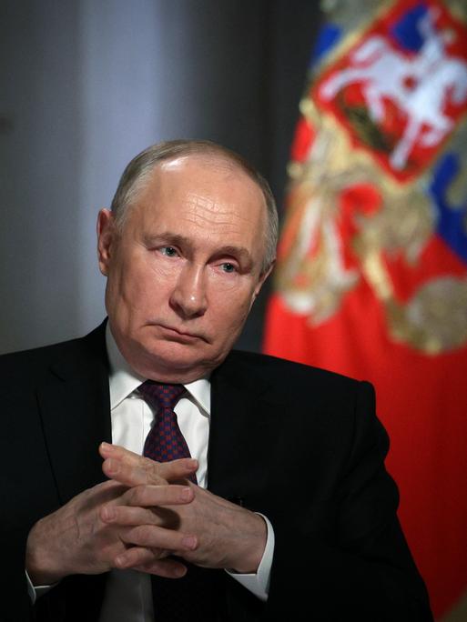Wladimir Putin sitzt während eines Interviews mit gefalteten Händen und ernstem Gesichtsausdruck dem Interviewer gegenüber.