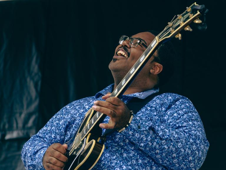 Ein Mann mit blauem Hemd und Gitarre steht auf einer Bühne und singt, während er den Kopf glücklich weit nach oben streckt.