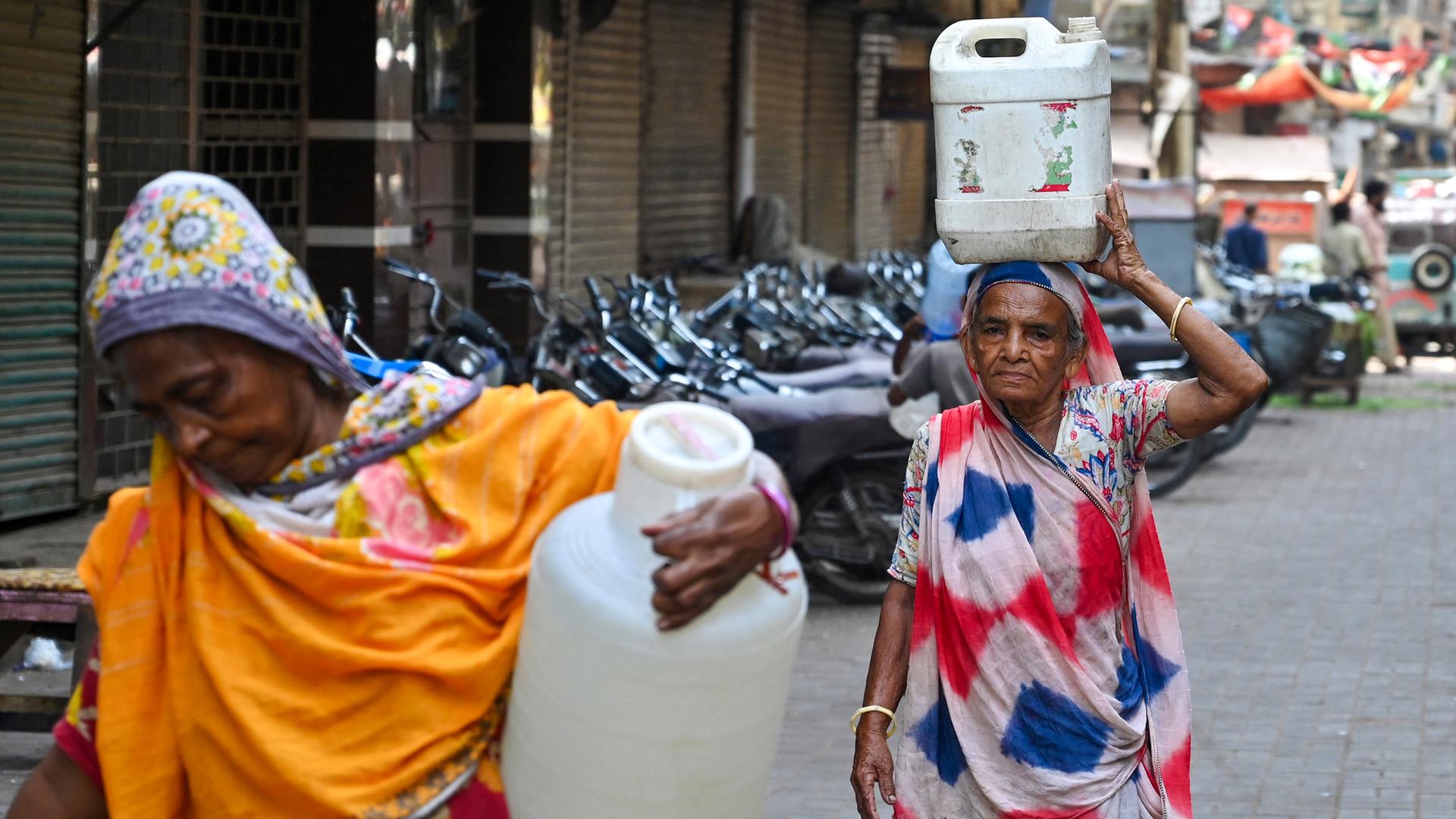 Zwei Frauen in Saris laufen mit schweren Wasserkanistern eine Straße entlang.