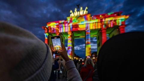 Das Brandenburger Tor wird nachts von bunten Lichtern angestrahlt. Im Vordergrund hält eine Person ein Handy hoch, um ein Foto zu machen. Das Brandenburger Tor wird nachts von bunten Lichtern angestrahlt. Im Vordergrund hält eine Person ein Handy hoch, um ein Foto zu machen.