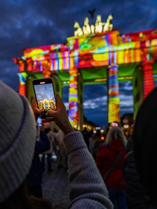 Das Brandenburger Tor wird nachts von bunten Lichtern angestrahlt. Im Vordergrund hält eine Person ein Handy hoch, um ein Foto zu machen. Das Brandenburger Tor wird nachts von bunten Lichtern angestrahlt. Im Vordergrund hält eine Person ein Handy hoch, um ein Foto zu machen.