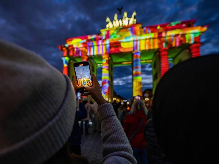 Das Brandenburger Tor wird nachts von bunten Lichtern angestrahlt. Im Vordergrund hält eine Person ein Handy hoch, um ein Foto zu machen. Das Brandenburger Tor wird nachts von bunten Lichtern angestrahlt. Im Vordergrund hält eine Person ein Handy hoch, um ein Foto zu machen.