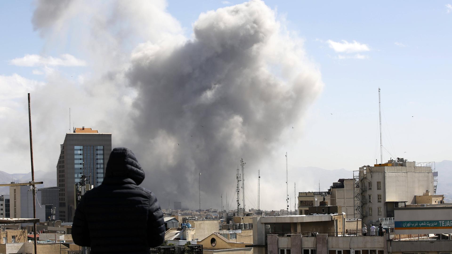 Angriff auf Teheran: Ein Mann schaut von einem Häuserdach über die Dächer der iranischen Hauptstadt. Im Hintergrund steigt nach einem Luftschlag Rauch auf. 