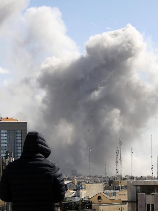 Angriff auf Teheran: Ein Mann schaut von einem Häuserdach über die Dächer der iranischen Hauptstadt. Im Hintergrund steigt nach einem Luftschlag Rauch auf. Angriff auf Teheran: Ein Mann schaut von einem Häuserdach über die Dächer der iranischen Hauptstadt. Im Hintergrund steigt nach einem Luftschlag Rauch auf.
