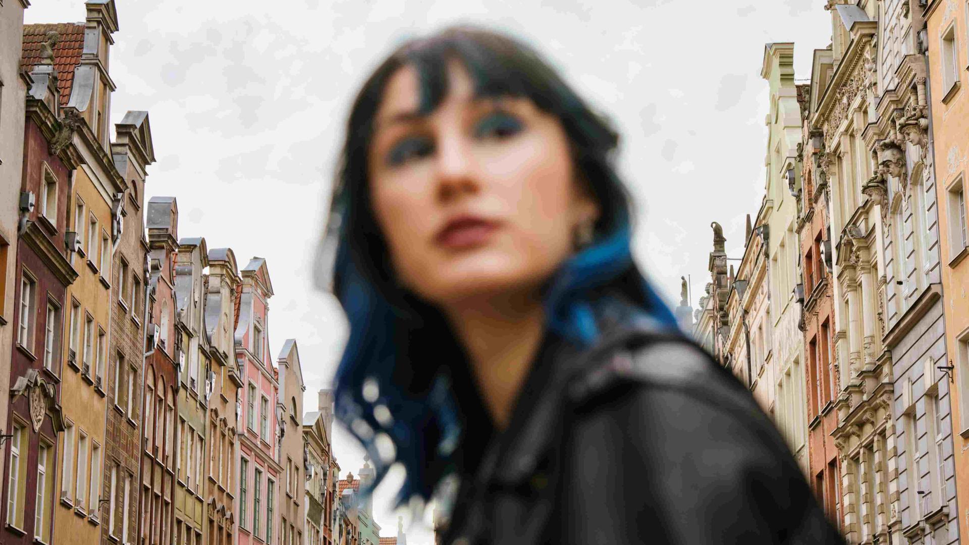 Unscharfes Porträt einer Frau mit blauen Haarsträhnen zwischen Altbaufassaden.