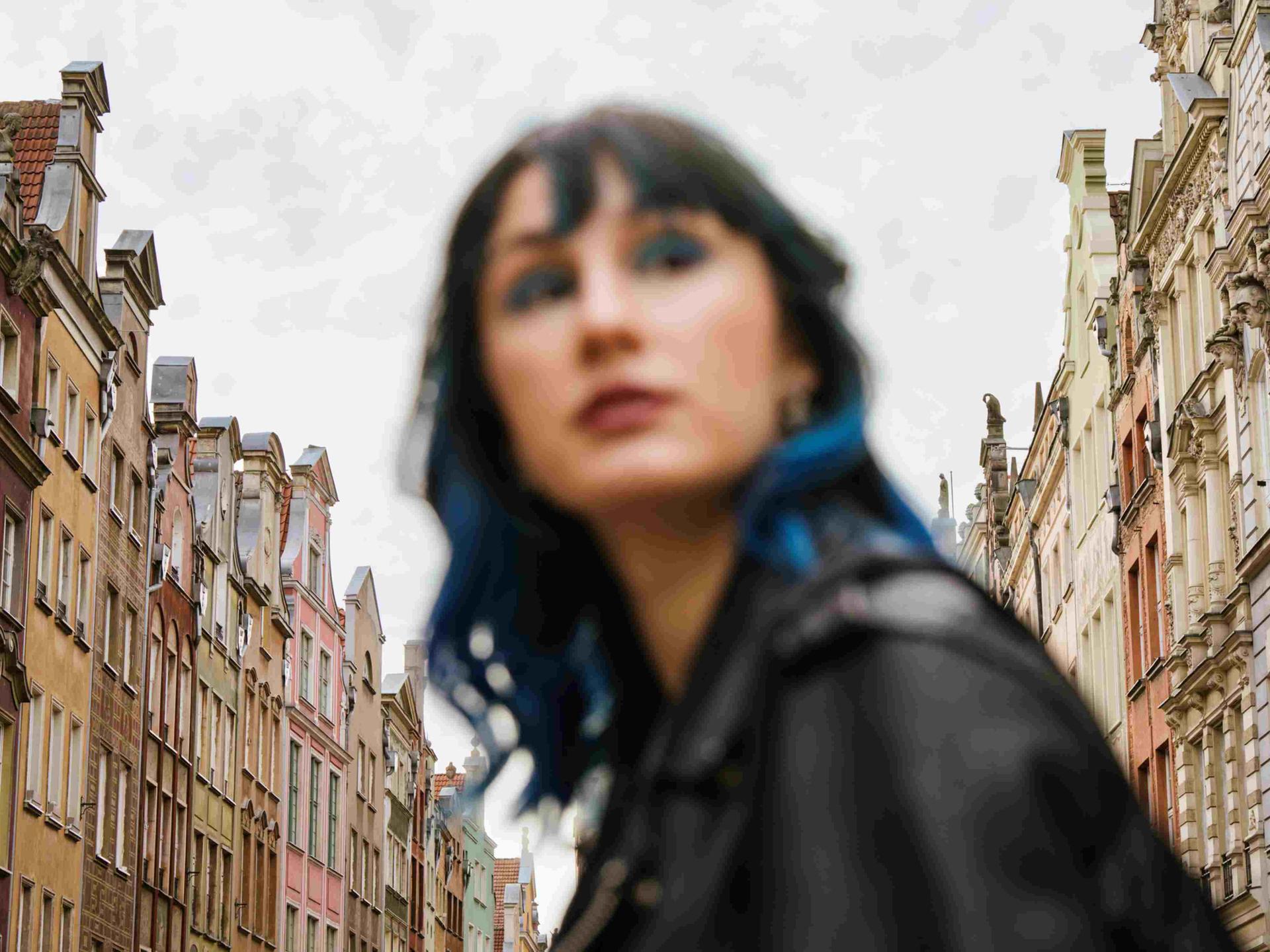 Unscharfes Porträt einer Frau mit blauen Haarsträhnen zwischen Altbaufassaden.