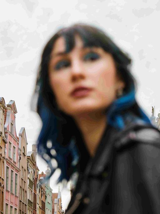 Unscharfes Porträt einer Frau mit blauen Haarsträhnen zwischen Altbaufassaden.
