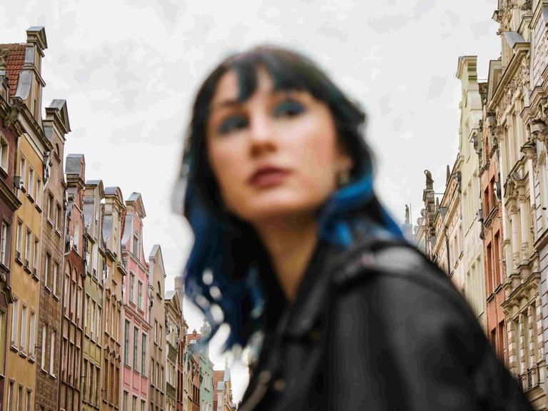Unscharfes Porträt einer Frau mit blauen Haarsträhnen zwischen Altbaufassaden.