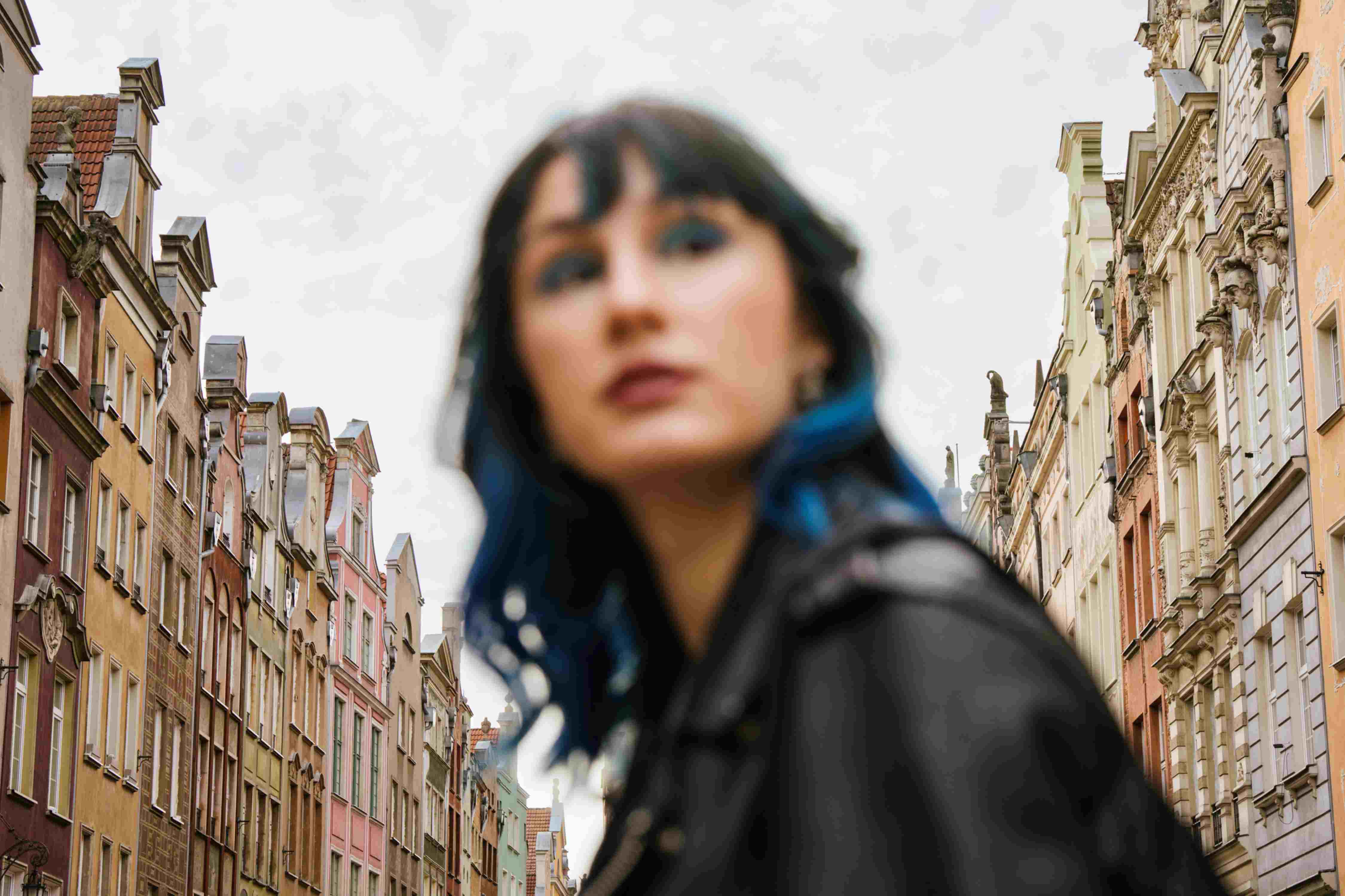 Unscharfes Porträt einer Frau mit blauen Haarsträhnen zwischen Altbaufassaden. Unscharfes Porträt einer Frau mit blauen Haarsträhnen zwischen Altbaufassaden.