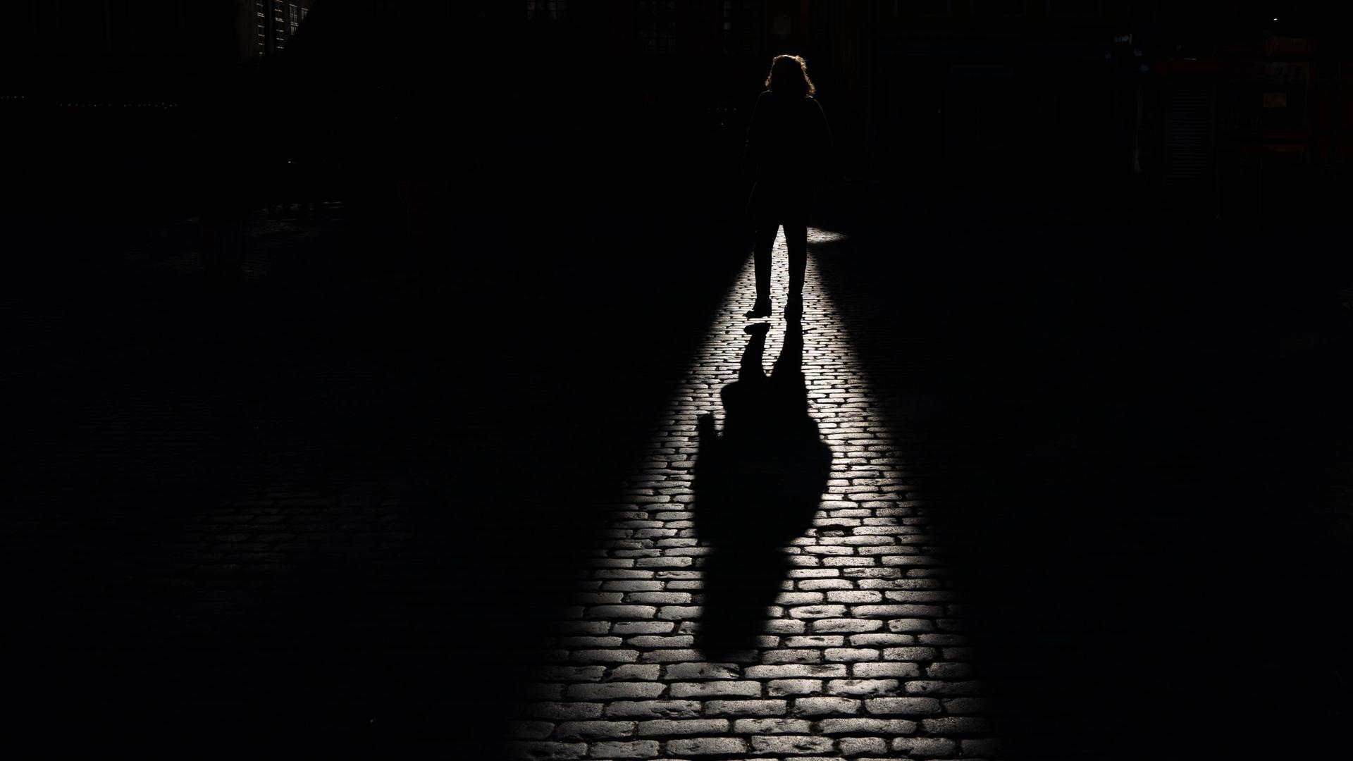 Schatten einer gehenden Person, der auf ein Kopfsteinpflaster fällt. Die Person selbst ist überwiegend im dunklen Bereich und ist nicht genauer zu erkennen. Schatten einer gehenden Person, der auf ein Kopfsteinpflaster fällt. Die Person selbst ist überwiegend im dunklen Bereich und ist nicht genauer zu erkennen.