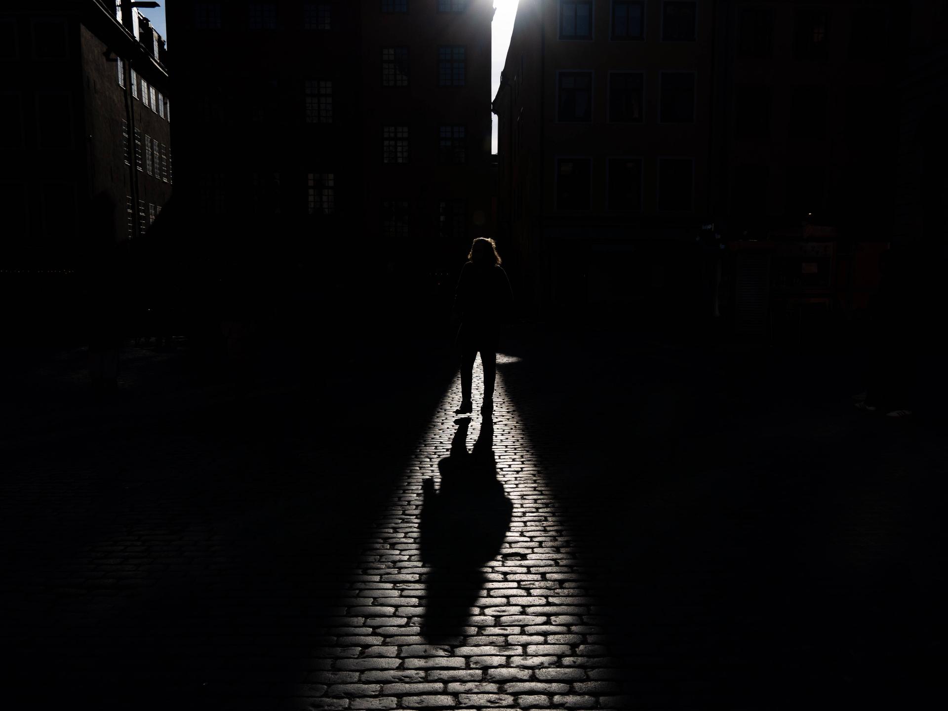 Schatten einer gehenden Person, der auf ein Kopfsteinpflaster fällt. Die Person selbst ist überwiegend im dunklen Bereich und ist nicht genauer zu erkennen. 