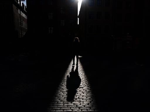 Schatten einer gehenden Person, der auf ein Kopfsteinpflaster fällt. Die Person selbst ist überwiegend im dunklen Bereich und ist nicht genauer zu erkennen. Schatten einer gehenden Person, der auf ein Kopfsteinpflaster fällt. Die Person selbst ist überwiegend im dunklen Bereich und ist nicht genauer zu erkennen.
