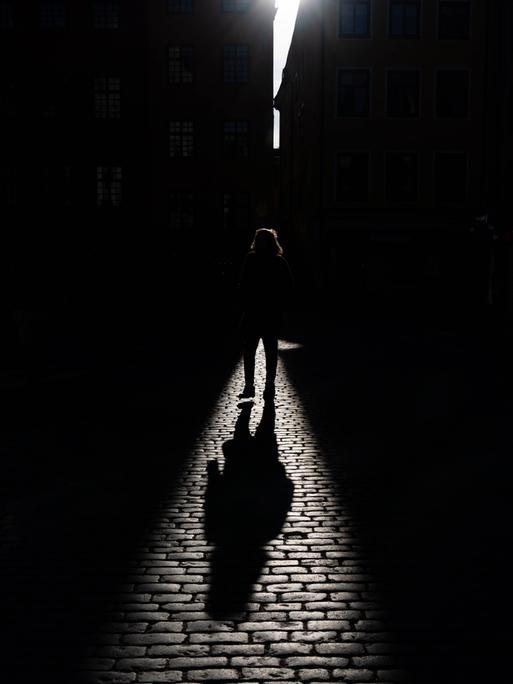 Schatten einer gehenden Person, der auf ein Kopfsteinpflaster fällt. Die Person selbst ist überwiegend im dunklen Bereich und ist nicht genauer zu erkennen. 