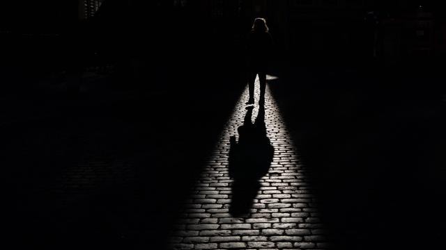 Schatten einer gehenden Person, der auf ein Kopfsteinpflaster fällt. Die Person selbst ist überwiegend im dunklen Bereich und ist nicht genauer zu erkennen. 