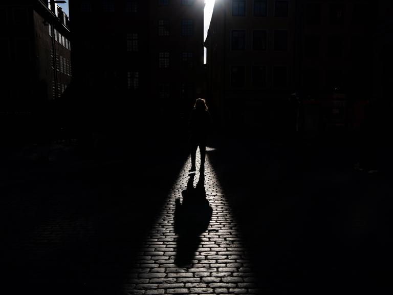 Schatten einer gehenden Person, der auf ein Kopfsteinpflaster fällt. Die Person selbst ist überwiegend im dunklen Bereich und ist nicht genauer zu erkennen. 