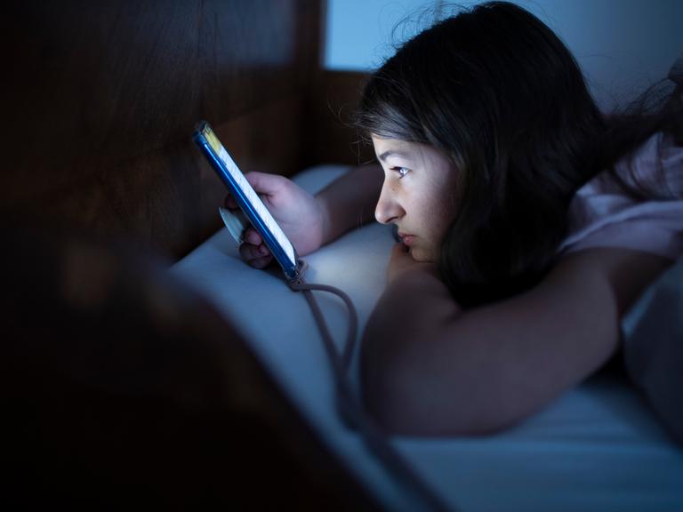 Ein Kind liegt mit einem Handy im Bett und schaut auf den Bildschirm.