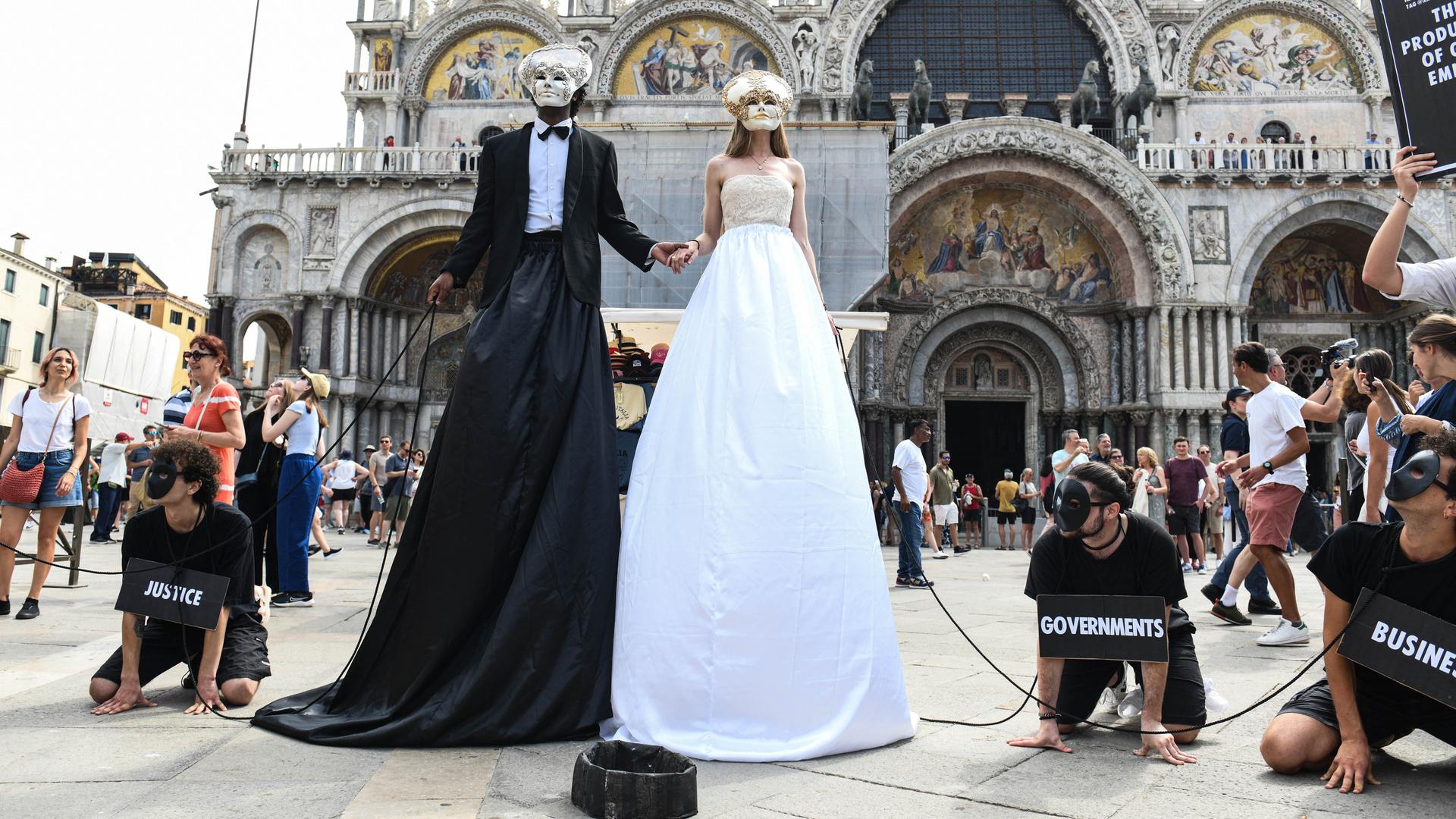 Zwei überhöhte Figuren, verkleidet als Brautpaar stehen am Markusplatz. Sie tragen venezianische Masken. Neben ihnen am Boden Männer auf allen Vieren, die sie an Ketten führen. Um den Hals tragen sie Schilder mit der Aufschrift "government" und "justice"