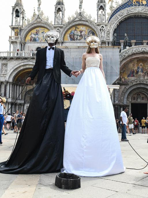 Zwei überhöhte Figuren, verkleidet als Brautpaar stehen am Markusplatz. Sie tragen venezianische Masken. Neben ihnen am Boden Männer auf allen Vieren, die sie an Ketten führen. Um den Hals tragen sie Schilder mit der Aufschrift "government" und "justice"