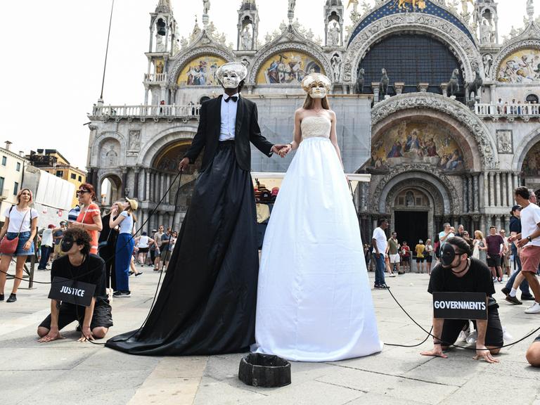 Zwei überhöhte Figuren, verkleidet als Brautpaar stehen am Markusplatz. Sie tragen venezianische Masken. Neben ihnen am Boden Männer auf allen Vieren, die sie an Ketten führen. Um den Hals tragen sie Schilder mit der Aufschrift "government" und "justice"