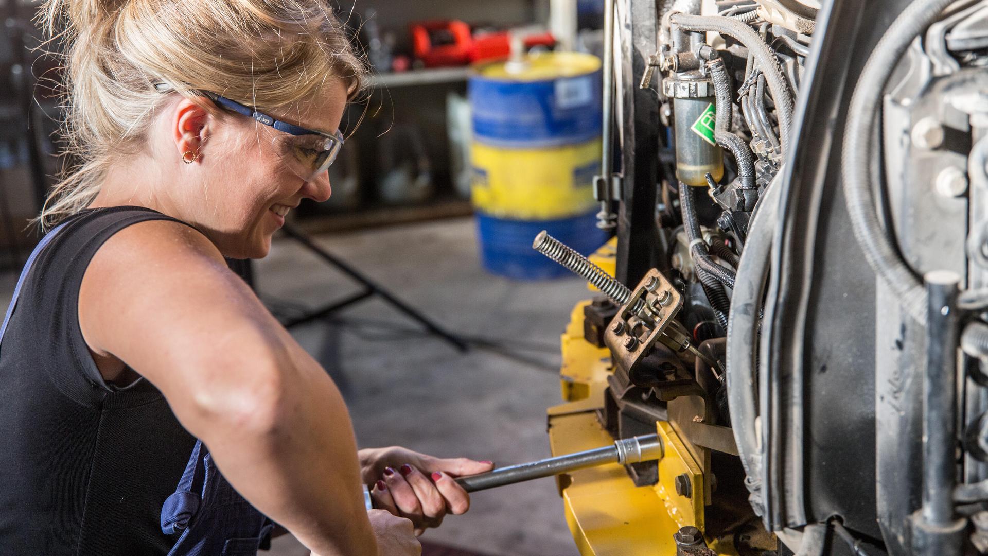 Eine Frau löst einem Knarrenschlüssel Verschraubungen und löst Teile. Bei der Arbeit trägt sie eine Schutzbrille und eine Latzhose.