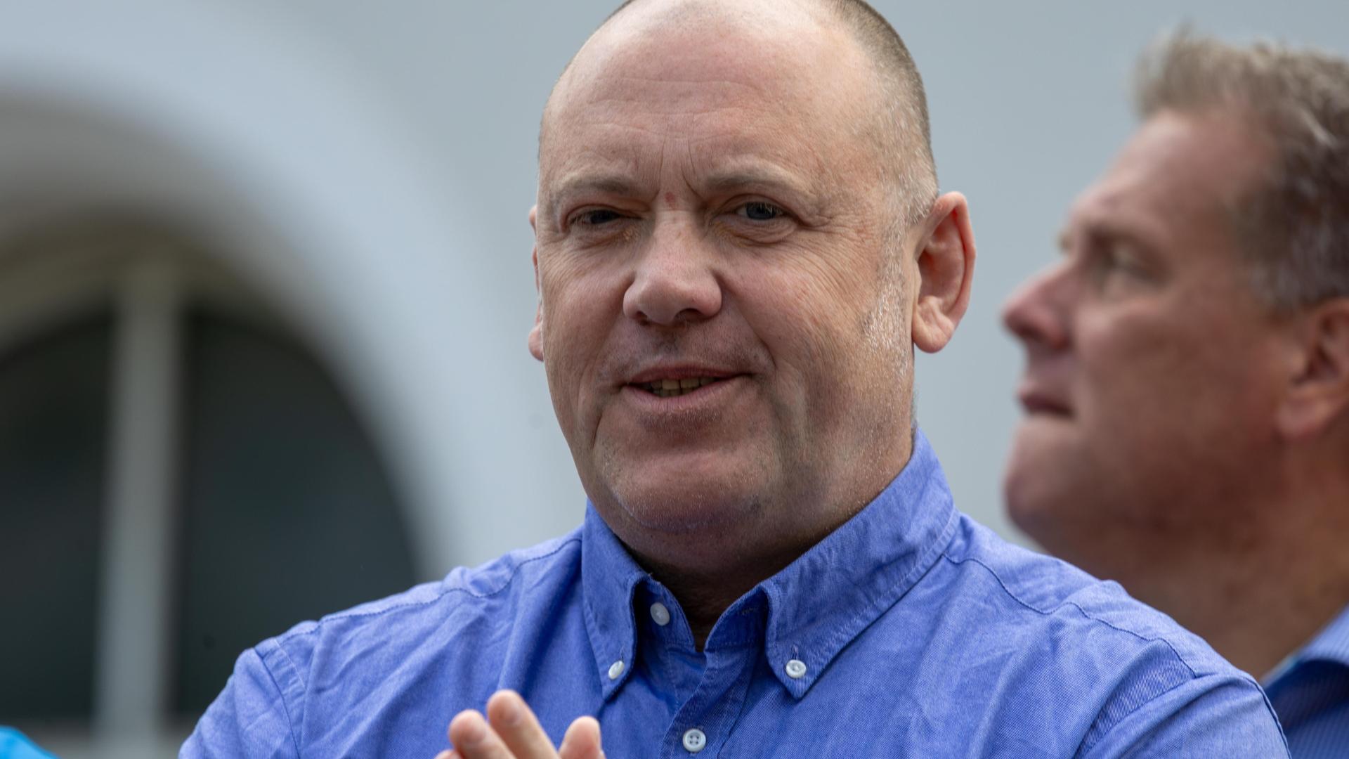  Paul im blauen Hemd. Er steht vor einem Gebäude, das man nur unscharf sieht, und lächelt. Dahinter ein weiterer Mann. 