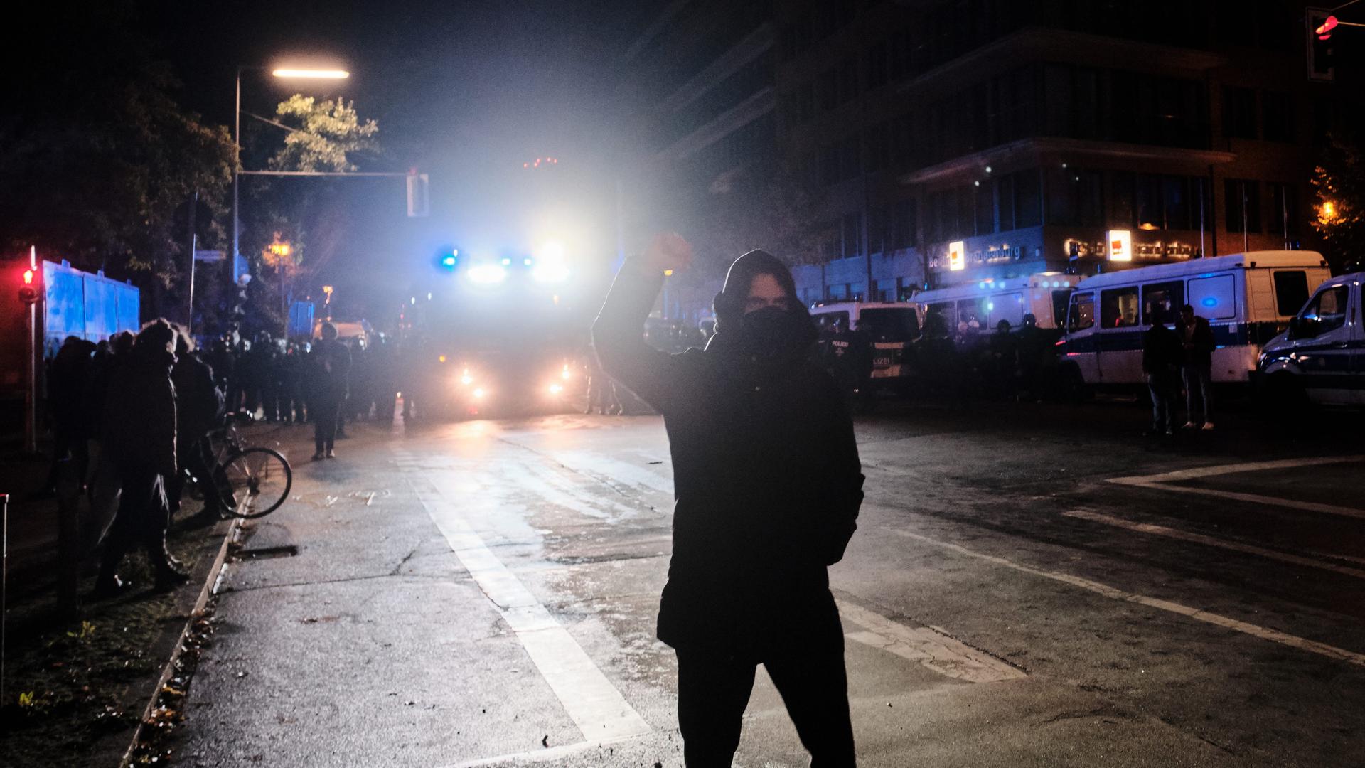 Eine schwarz gekleidete und maskierte Person steht während einer Demonstration im Scheinwerferlicht eines Polizeifahrzeugs und streckt kämpferisch die Faust in die Höhe.