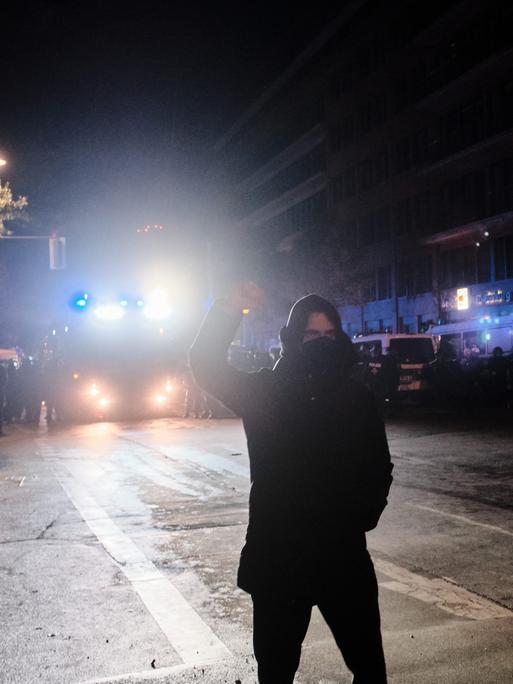 Eine schwarz gekleidete und maskierte Person steht während einer Demonstration im Scheinwerferlicht eines Polizeifahrzeugs und streckt kämpferisch die Faust in die Höhe.