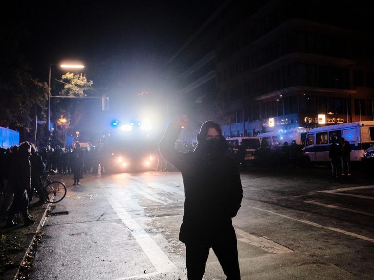 Eine schwarz gekleidete und maskierte Person steht während einer Demonstration im Scheinwerferlicht eines Polizeifahrzeugs und streckt kämpferisch die Faust in die Höhe.