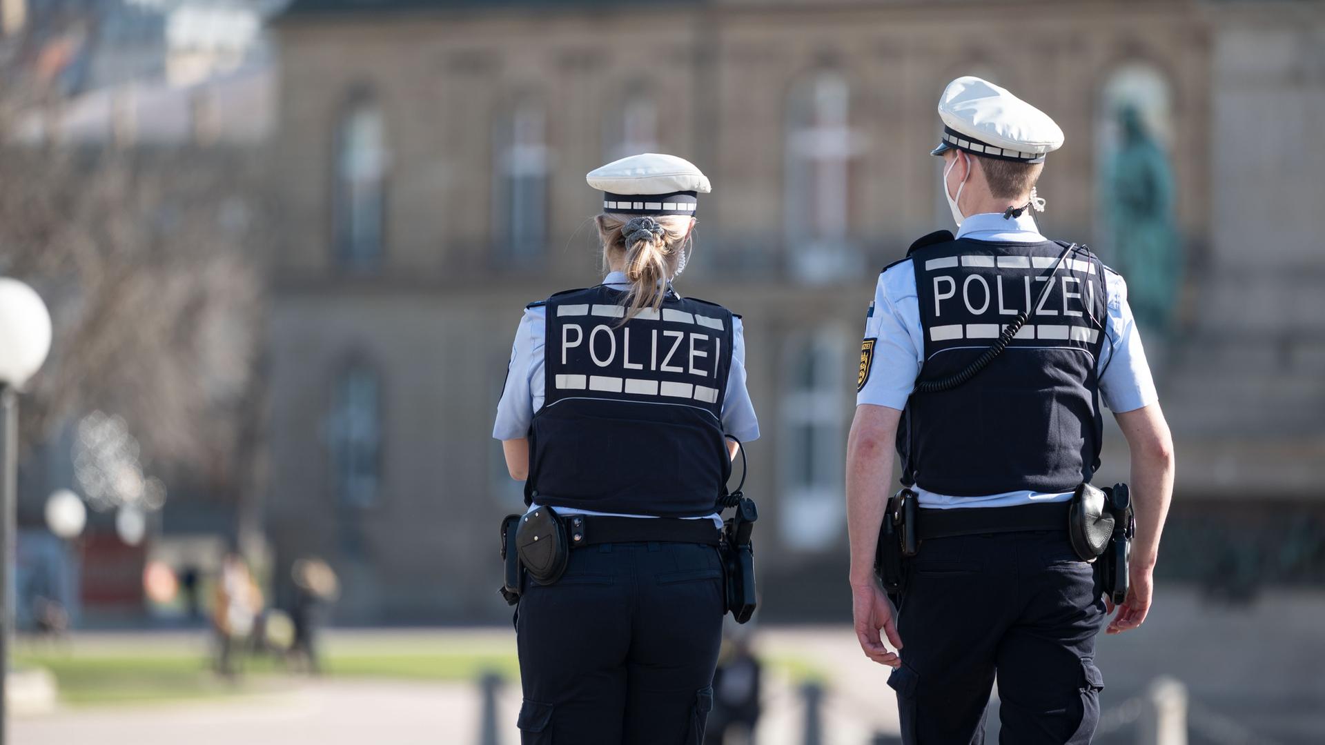 Ein Polizist und eine Polizistin gehen über den Schlossplatz in Stuttgart, das Schloss ist im Hintergrund zu sehen. Sie haben dem Betrachter den Rücken zugewandt.