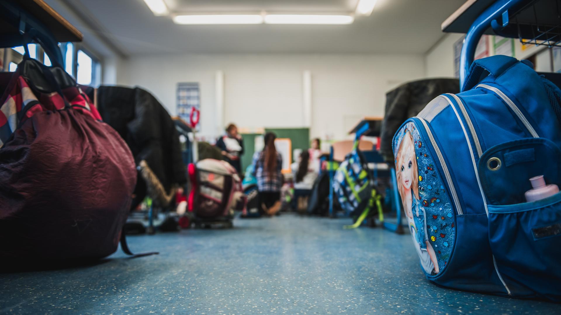 Blick in einen Klassenraum, wo an den Schultischen Schultaschen stehen und Schüler sitzen und stehen.