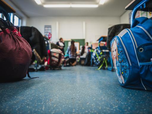 Blick in einen Klassenraum, wo an den Schultischen Schultaschen stehen und Schüler sitzen und stehen.