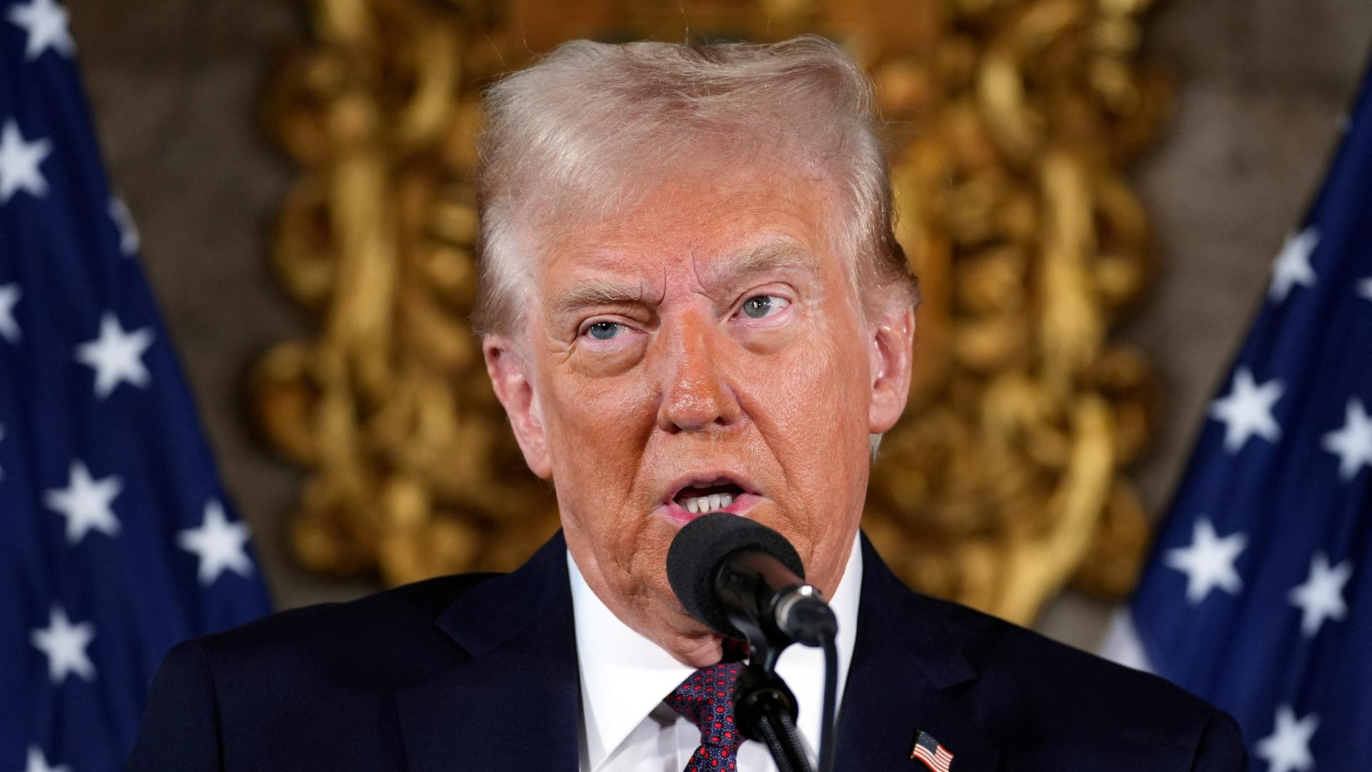 Der künftige US-Präsident Donald Trump bei einer Pressekonferenz in Florida. Er spricht in ein Mikrophon. Im Hintergrund sind US-Flaggen zu sehen. 