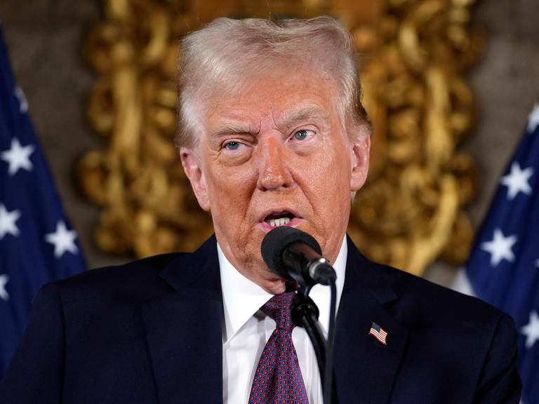 Der künftige US-Präsident Donald Trump bei einer Pressekonferenz in Florida. Er spricht in ein Mikrophon. Im Hintergrund sind US-Flaggen zu sehen.  