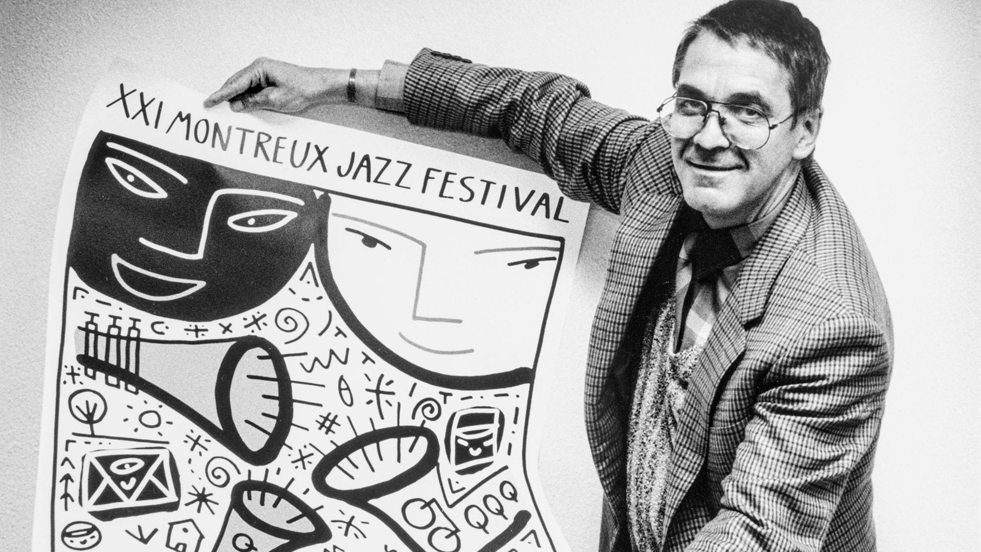 Claude Nobs hält auf dem Schwarz.Weiß-Bild das Plakat zum 21. Montreux Jazz Festival in den Händen und zeigt es in die Kamera. Er trägt das Haar kurz und hat eine Brille.