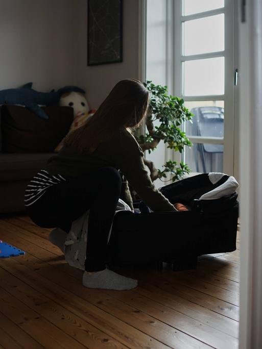 Eine Frau in einer Altbauwohnung beugt sich über eine Babytragetasche.